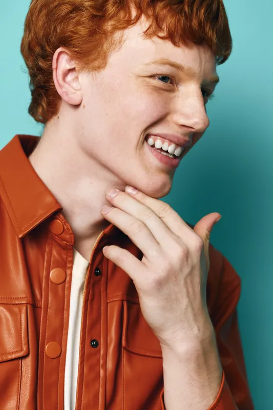 Young man with red hair smiling and touching his face wearing a brown leather jacket against a blue background. Portrait, happiness, casual style, and natural expression.
