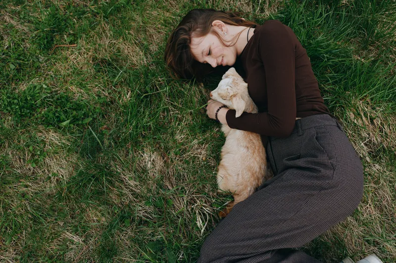Woman lies on grass outdoors, hugging a small goat kid, calm and affectionate, wearing a brown sweater and long skirt, portraying a peaceful lifestyle and intimate bond with nature.