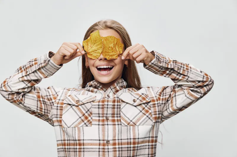 Girl smiling and holding two autumn leaves in front of her eyes wearing plaid shirt with long hair isolated on white background for fall nature concept and happy child.