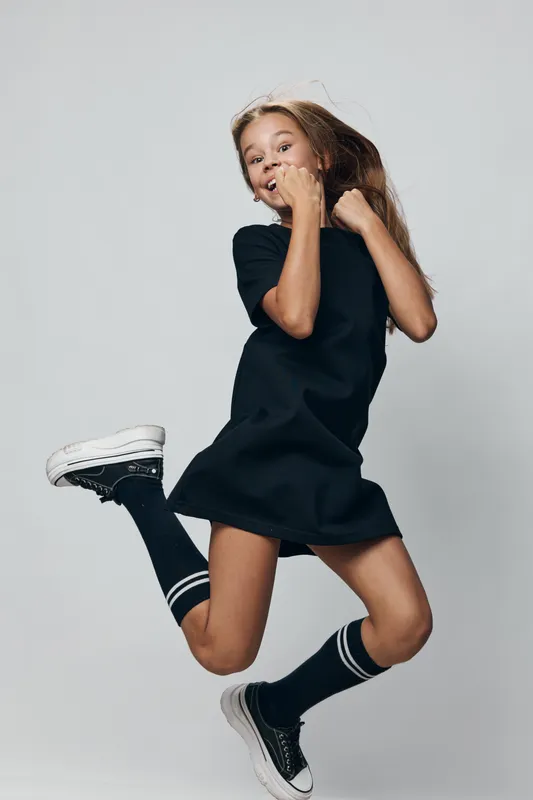 Girl jumping in casual black dress and sneakers with knee-high socks on light background. Active child with hair movement in joyful motion for youth and energy.