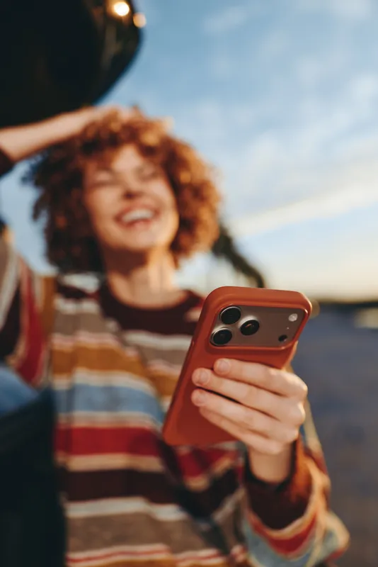 Woman lifestyle smartphone selfie, curly hair outdoors sunset casual striped sweater, holding phone with warm light, soft bokeh background, relaxed candid everyday mood, copy space