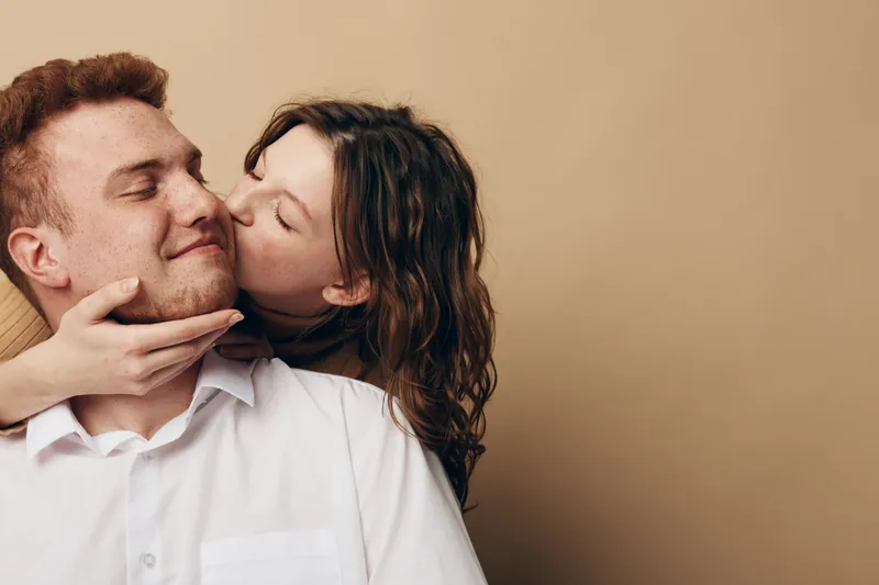 Young woman kisses smiling man on cheek while holding his face gently. Intimate moment of affection and love between couple on beige background. Concept of romance and tenderness.