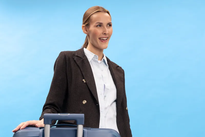 Smiling woman in business attire stands with suitcase on blue background. Concept of travel, business trip, professional lifestyle, and positive attitude in modern work environment.