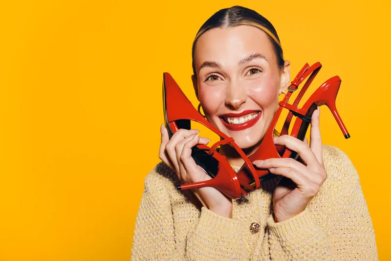 Happy woman with red lipstick holds red high heel shoes near face smiling on yellow background. Concept of fashion, style, shopping, and joyful emotions in studio lighting.