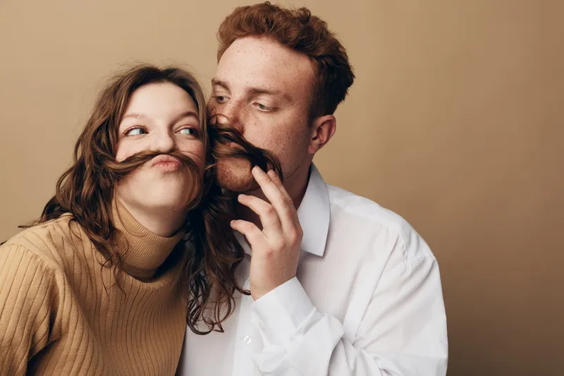 Playful young couple with brown hair making mustaches from hair, smiling and having fun together in casual clothes on beige background. Concept of love, friendship, and joyful moments.