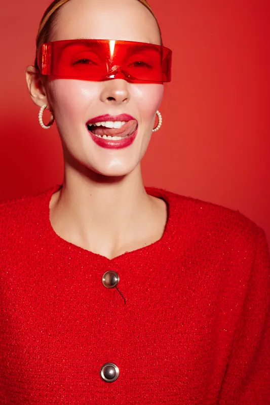 Young woman in red sweater and red futuristic glasses smiles with tongue out on red background. Concept of fashion, style, modern accessories, and playful attitude.