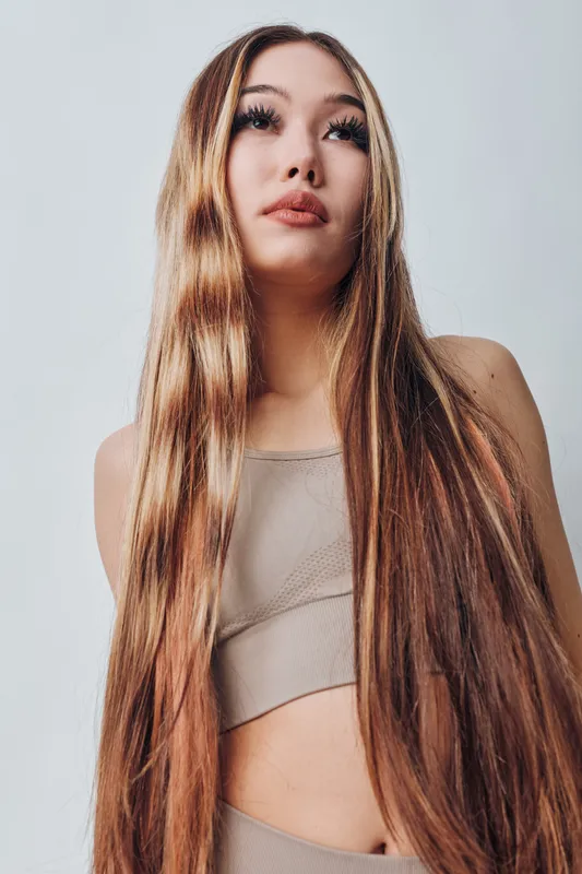 Portrait of woman with long hair wearing beige top standing against white background looking up with natural makeup and serious expression in studio lighting.