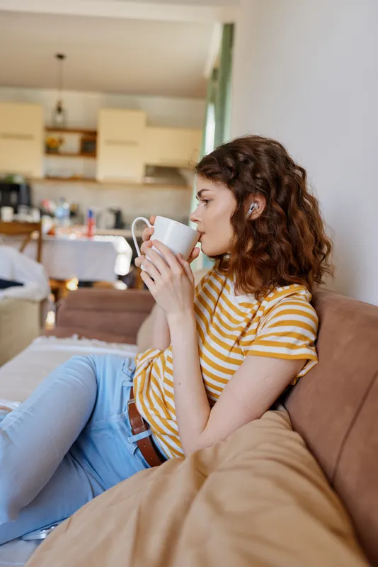 Woman home casual relaxed sitting mug coffee couch, enjoying sunlight indoors, cozy moment with soft ambience, lifestyle focused on comfort and everyday leisure. Bright, airy vibe, natural tones.
