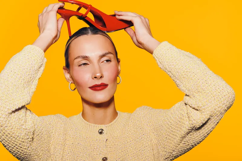 Young woman with red lipstick and hoop earrings holds red high heel shoes above her head against bright yellow background. Concept for fashion, style, and confident attitude.