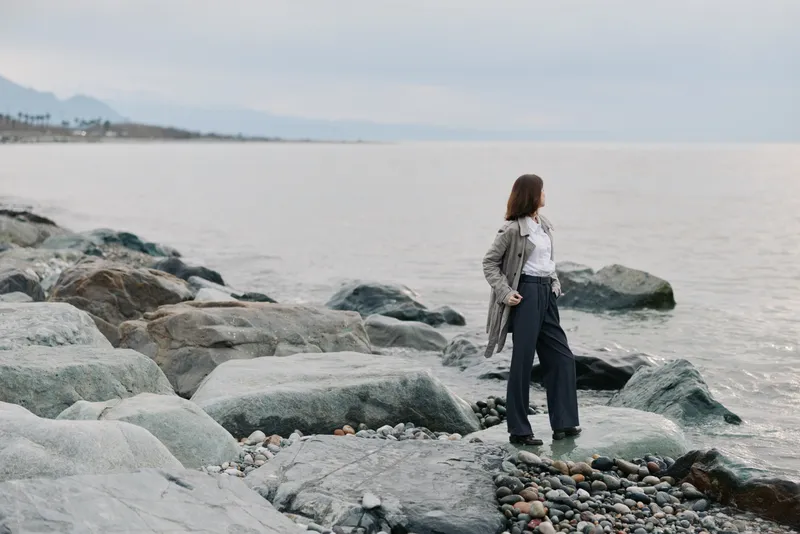 Woman in casual clothes stands on rocky shore looking at calm sea under cloudy sky. Concept of solitude, nature, and peaceful coastal landscape with soft natural light and relaxed mood.