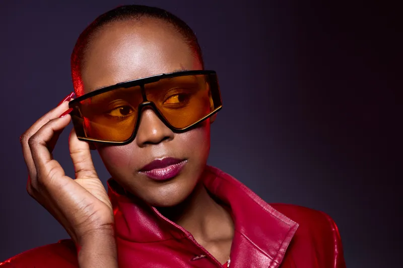 Close-up of confident African woman with dark skin wearing oversized orange sunglasses and red leather jacket. Studio portrait on dark background with stylish fashion and attitude concept.