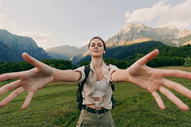 Young woman with long hair wearing casual outdoor clothes reaches her arms forward in a green mountain valley under a partly cloudy sky expressing freedom and adventure.
