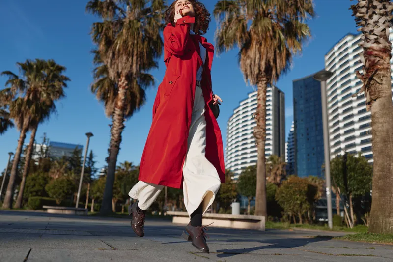 Young woman in red coat walking outdoors on a sunny day with palm trees and modern buildings in the background, urban lifestyle and fashion concept.