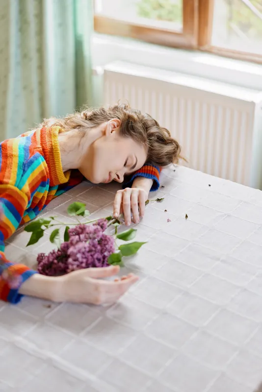 A tired woman lies across a table in a cozy home, head resting on her arm near a bouquet of flowers. A quiet moment capturing fatigue, exhaustion, and the need to pause.