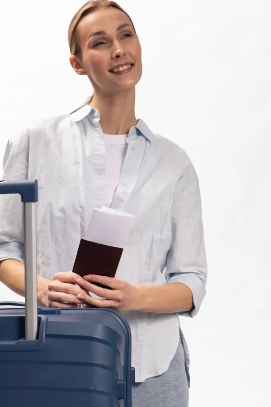 Smiling woman traveler holds passport and boarding pass near blue suitcase. Casual clothing and relaxed expression. Concept for travel, vacation, tourism, and journey planning.