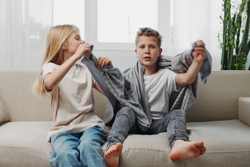Two children sitting on a beige sofa, a girl helping a boy put on a grey hoodie, casual home setting with natural light. Concept of childhood, family, and everyday moments.