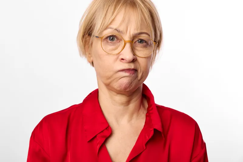 Senior woman with glasses looks worried and serious, head and shoulders portrait on a white background, visible stress and concern on her face, studio concept