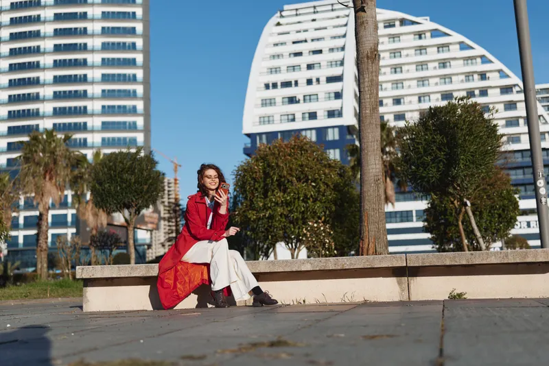 Young woman sitting outdoors on a stone bench talking on smartphone, casual outfit, sunny day, modern buildings and greenery in background
