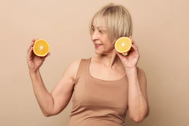 Smiling mature woman with short blonde hair holding two orange halves near face on beige background. Concept of healthy lifestyle, nutrition, and natural vitamins for wellbeing.