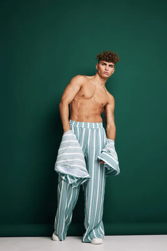 Young shirtless man with curly hair wearing loose striped pants stands confidently against dark green background holding striped shirt in his hands. Fashion pose and casual style.