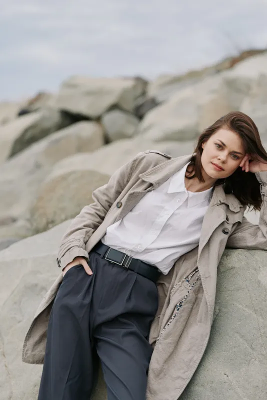 Young woman in beige coat and white shirt leans on rocks outdoors. Casual style with relaxed pose, natural makeup, and soft lighting. Concept of fashion, nature, and calm mood.