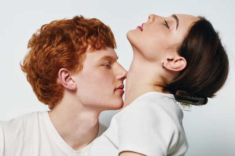 Couple with red hair man and woman with dark hair in intimate pose on white background. Young lovers, tenderness and connection in studio portrait.