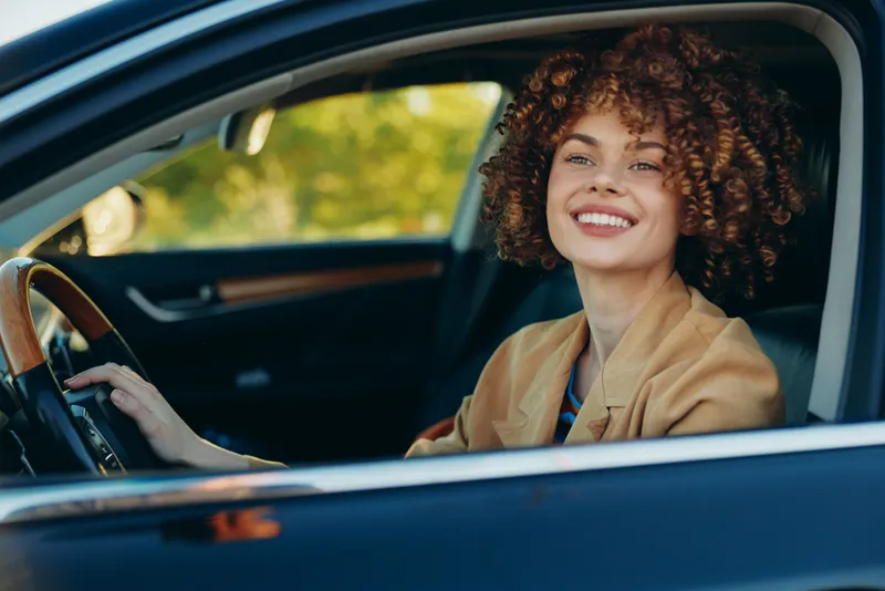 Happy woman driving a car, smiling at the road, casual blazer, retro film color grading, lifestyle moment, focused on journey, daylight on a quiet street, candid and upbeat