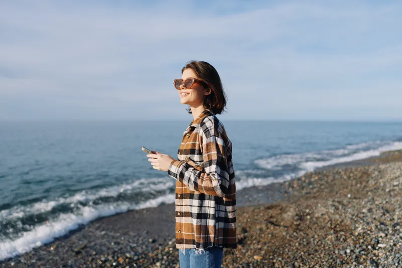 Woman standing on a pebbled beach with smartphone, wearing a plaid shirt and sunglasses, enjoying sunny weather and sea breeze beside the coast in a relaxed pose