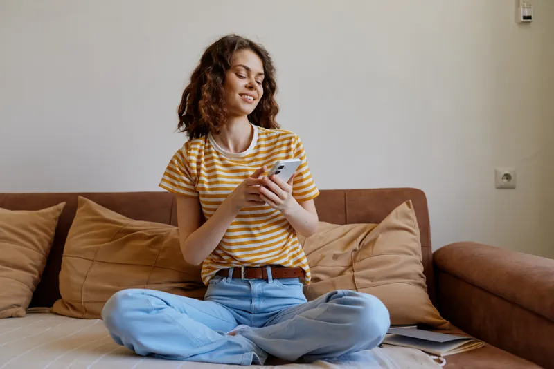 A relaxed woman sits crosslegged on a sofa at home, smiling while scrolling her phone. A casual lifestyle scene focused on comfort, quiet moments, and everyday connectivity.