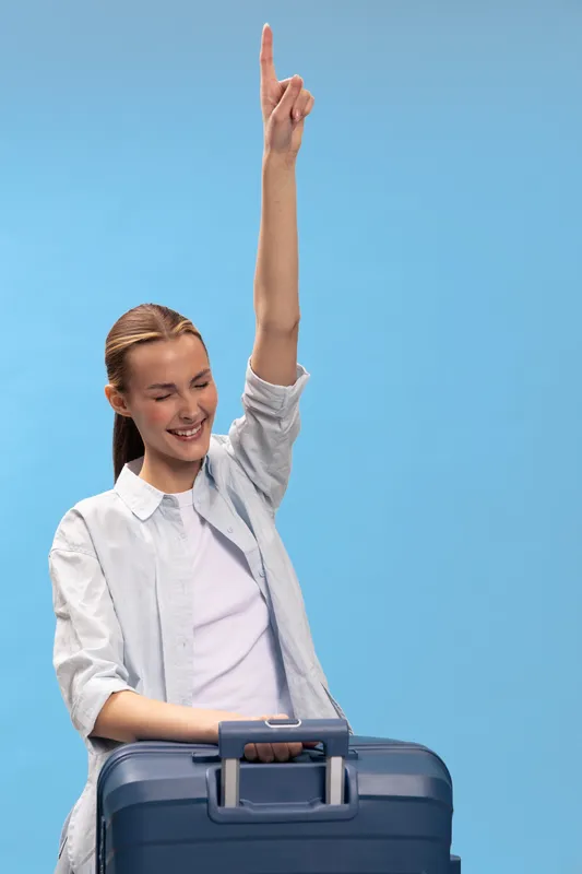 Young woman with suitcase raises hand and smiles standing against blue background. Concept for travel, vacation, joy, and freedom with casual outfit and happy mood.