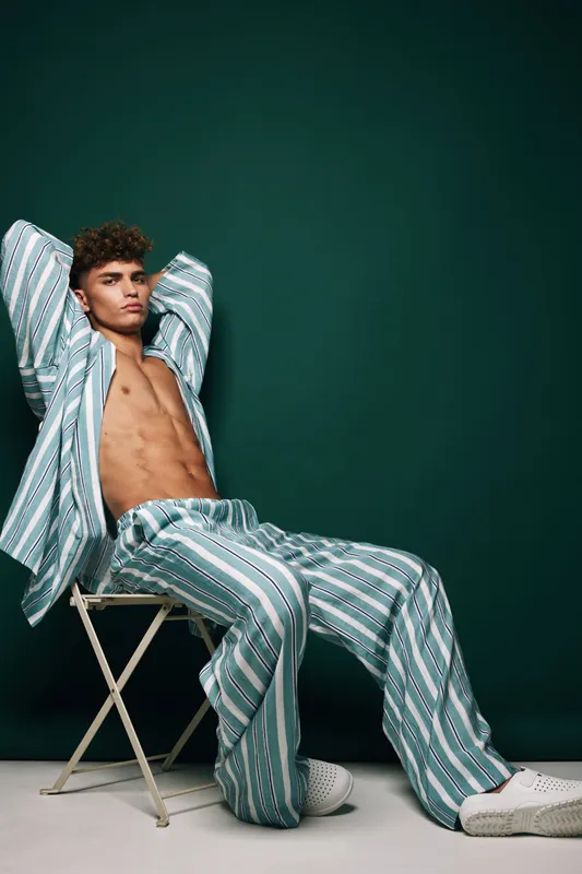 Young man in striped outfit leaning back on chair posing confidently against dark green background indoors fashion and style portrait of male model lounge look