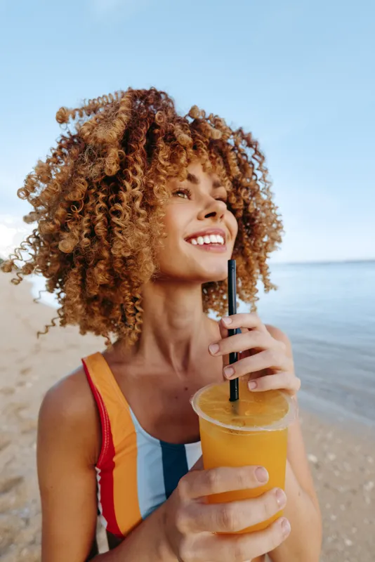 Woman with curly hair holding juice with a black straw on a sunny beach, smiling and looking at the ocean, summer vacation mood, refreshing drink, bright natural light, casual portrait