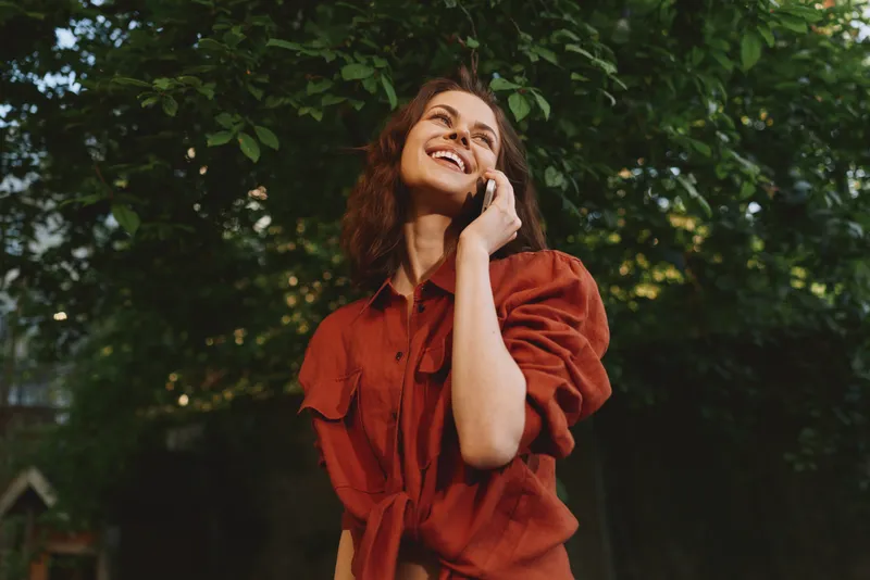 Cheerful woman outdoors wearing a red shirt, smiling with eyes closed, sunlit moment in lush greenery, relaxed posture, natural colors, candid lifestyle portrait