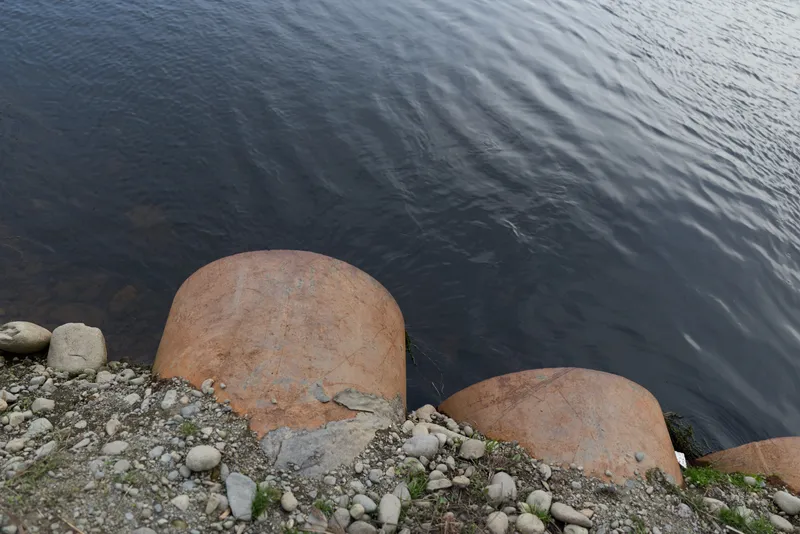 River shoreline with clear water, rounded rocks and rust colored boulders, scattered pebbles on bank, outdoor nature scene with calm flow, natural texture, peaceful mood, soft light