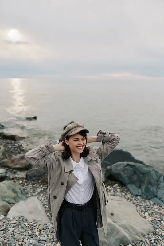 Young woman in stylish coat and cap smiles happily standing on rocky beach near calm sea under cloudy sky. Concept of fashion, nature, relaxation, and outdoor lifestyle by the water.