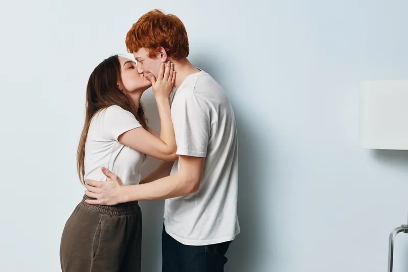 Couple kissing and hugging indoors, young woman and man standing close with eyes closed, affection and love in casual clothes against plain background.