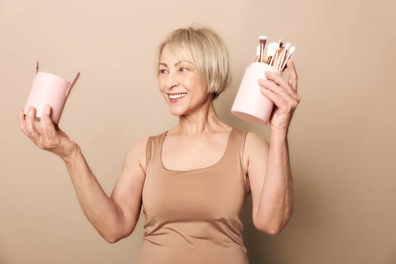 Smiling mature woman with short blonde hair holds two pink containers with makeup brushes in beige studio. Concept for beauty routine, cosmetics tools, self-care, and confident aging.
