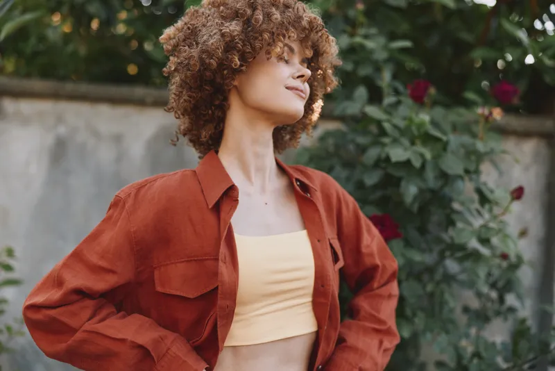 Woman lifestyle portrait outdoors, casual fashion, curly hair, warm jacket and crop top, relaxed pose in garden, conveying confidence, joy, and everyday elegance
