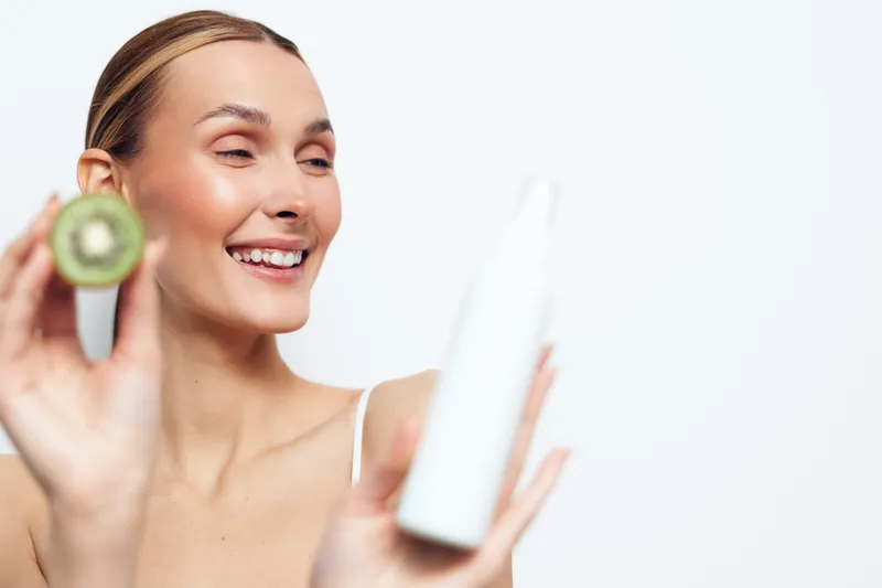 Smiling woman holding kiwi slice and white bottle skincare product with natural ingredients isolated on white background, enjoying healthy beauty routine and fresh healthy lifestyle