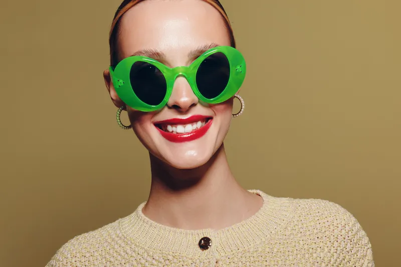 Smiling woman with green sunglasses and red lipstick wears beige sweater on brown background. Concept of fashion, style, beauty, and confident female portrait in studio lighting.