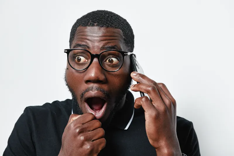 African American man with dark skin and glasses talking on phone with surprised expression, wearing black polo shirt on white background. Communication, emotion and technology concept.