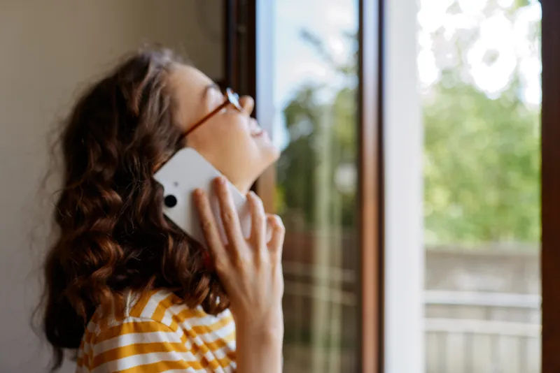 Young woman with curly hair and glasses chats on a phone by a bright window, enjoying a casual home moment in a sunlit interior. Her striped shirt complements the warm, relaxed atmosphere as she