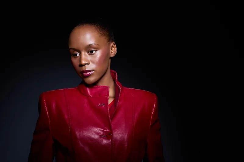 Elegant dark skinned African woman in red jacket poses against black background. Studio portrait with dramatic lighting highlights her confident expression and fashion style.