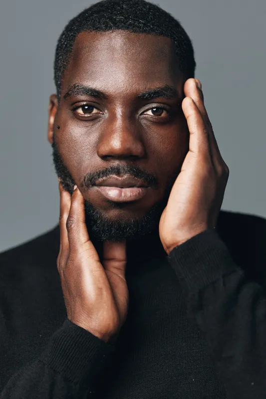 African man with dark skin and beard posing with hands on face in creative conceptual visual style. Closeup portrait with serious expression and black sweater on neutral background.