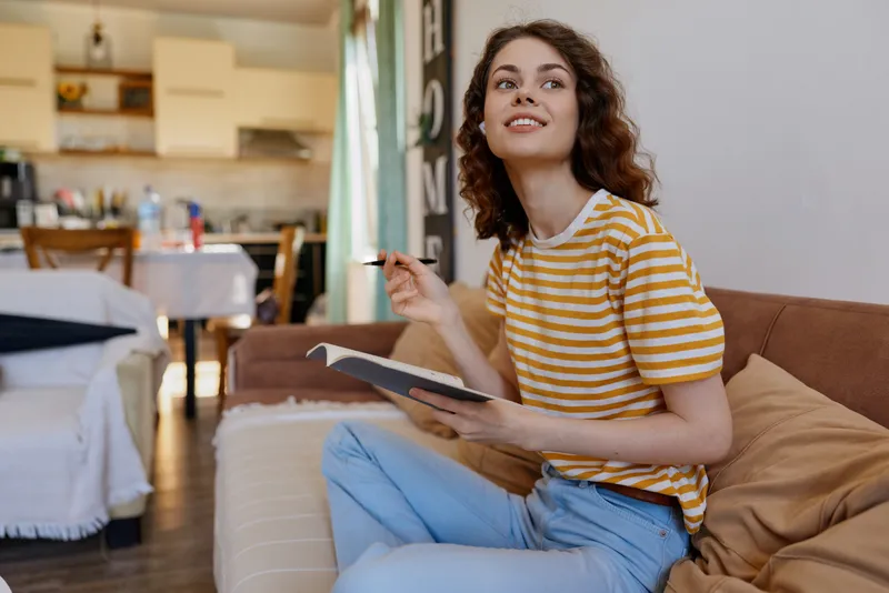 Woman at home on a sofa, casual striped shirt, writing in a notebook while using a tablet, relaxed mood, bright sunlight, cozy interior, everyday lifestyle. Creative routine and warm setting.