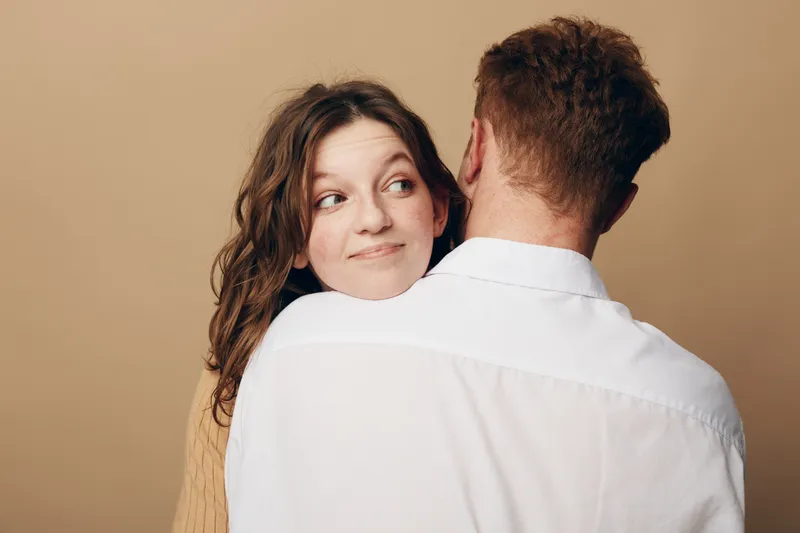 Young woman with curly hair embraces man wearing white shirt, looking sideways with smile. Intimate moment, love, relationship, and affection concept on beige background.