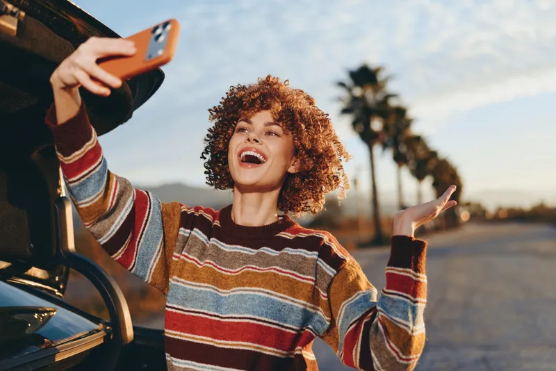 Woman lifestyle outdoors, smiling with curly hair, laughing while holding smartphone for selfie near car open door, warm sunset sunlight, relaxed sweater, palm lined street background