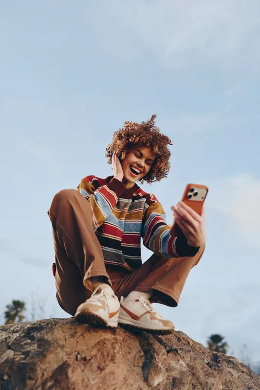 Smiling woman taking a selfie outdoors, casual lifestyle portrait while sitting on a rock under blue sky, colorful sweater and sneakers, natural light, travel mood with phone camera in summer