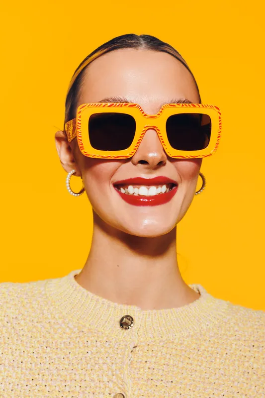 Smiling woman with bright red lipstick wears large orange sunglasses and yellow sweater against vibrant yellow background. Concept of fashion, summer style, and confident beauty.