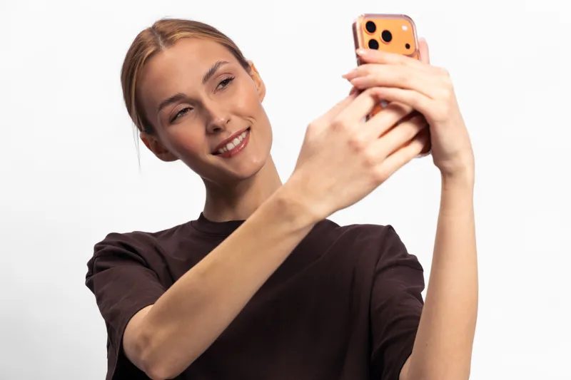 Young woman with light skin and red hair smiles while taking a selfie on a smartphone in a studio with a white background. Concept of social media, technology, and self-expression.